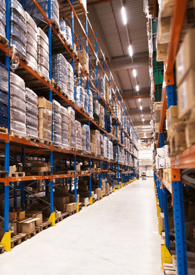 A long aisle in a warehouse with tall shelves on both sides, stacked with various boxes and courier packages.